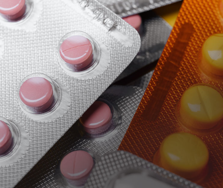 Close-up of various blister packs containing round pills; pink pills in metallic silver packaging and yellow pills in orange packaging. The packs overlap each other.