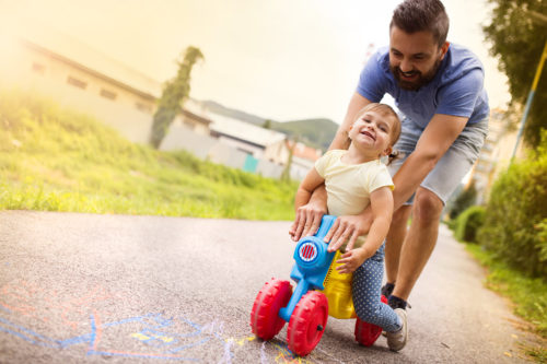 A man joyfully helps a smiling toddler ride a colorful plastic tricycle on a sunny path. Chalk drawings brighten the ground, evoking the carefree spirit often overshadowed by concerns like daycare abuse. Greenery and buildings frame this moment of innocence in the background.