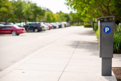 A parking lot with multiple parked cars and a sidewalk graced by trees. A parking meter with a blue "P" sign stands in the foreground, marking this peaceful day free from any parking lot accident under the clear, sunny sky.