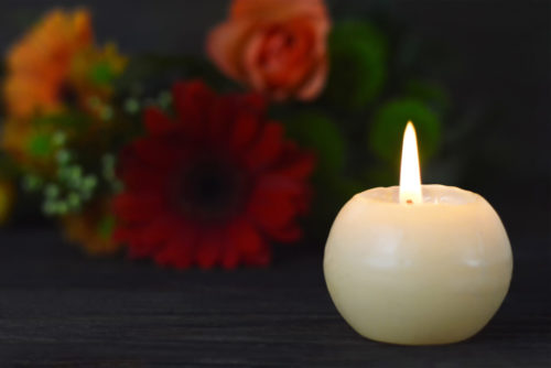 A round white candle is lit on a dark surface. In the blurred background, colorful flowers in shades of red and orange are visible, softly hinting at solace amidst life's wrongful death. The scene conveys a warm, peaceful ambiance.