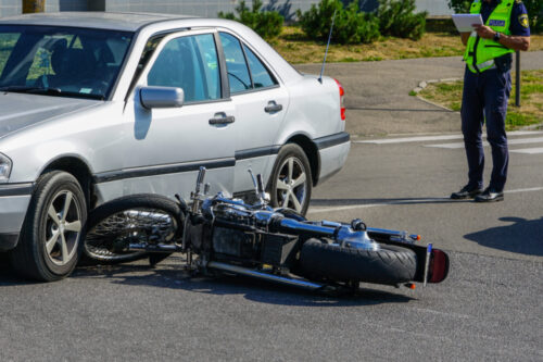 A silver car is embroiled in a motorcycle accident, with a black bike lying on its side in the road. A police officer stands nearby taking notes under the sunny sky of an urban area.