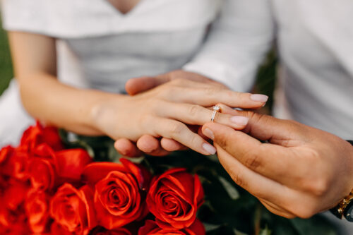 A person places a ring on another's finger, with a bouquet of red roses in the foreground. Both individuals wear white clothing, suggesting a romantic or engagement moment.